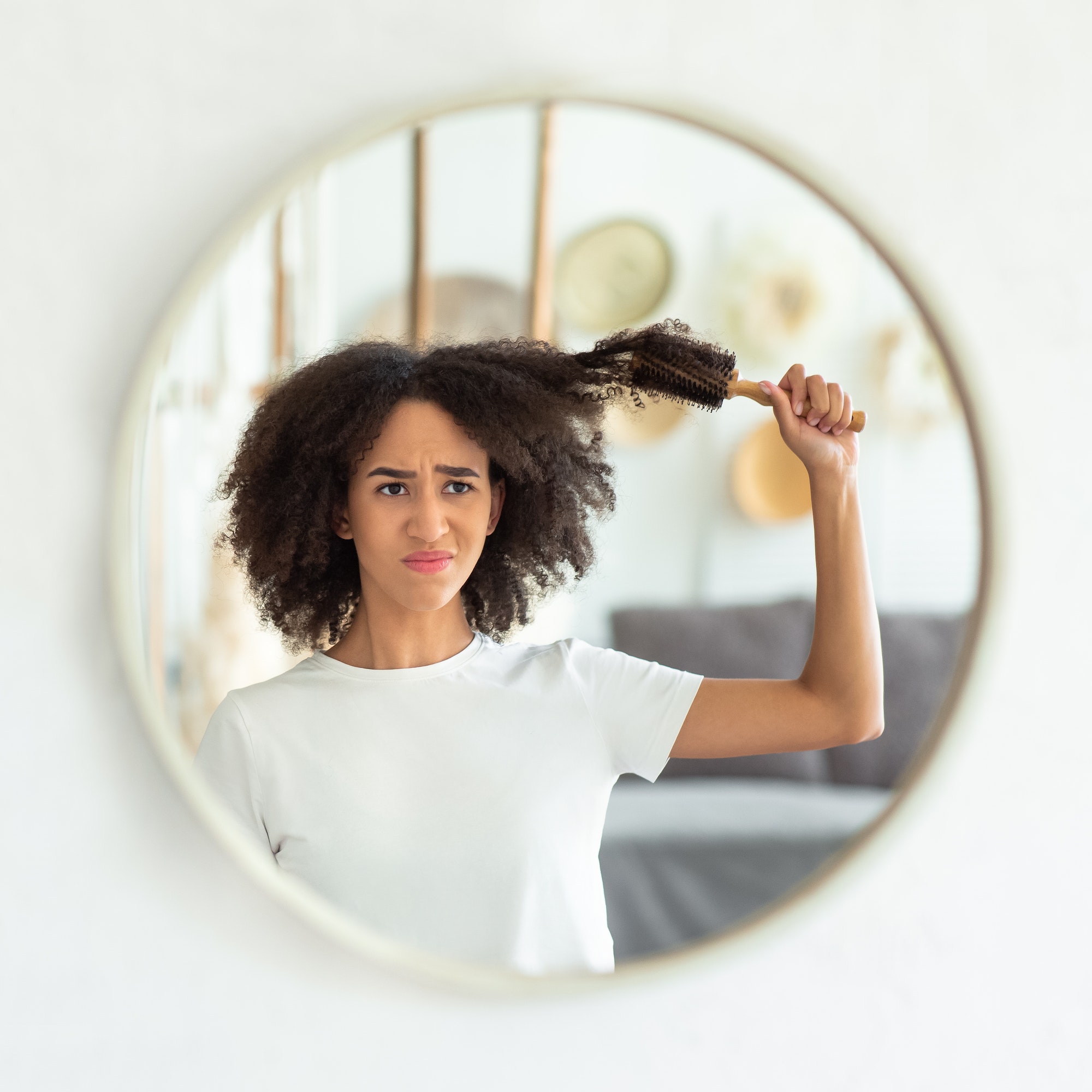 Upset young attractive african american lady pulls out comb from curly hair, looks in reflection of